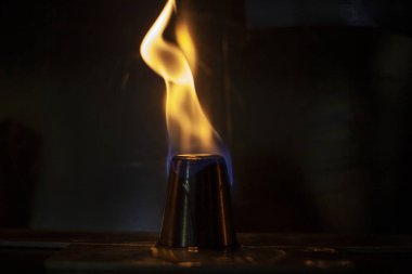 Alkol, alkollü içecekler için yığınla yakar. Metal bir bardağın etrafında alev dilleri. Ateşin dokusu. Siyah arka planda sarı ateş. Barda yangın çıktı. Alkolü ateşe verme geleneği..