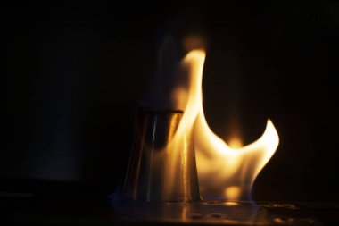 Alkol, alkollü içecekler için yığınla yakar. Metal bir bardağın etrafında alev dilleri. Ateşin dokusu. Siyah arka planda sarı ateş. Barda yangın çıktı. Alkolü ateşe verme geleneği..