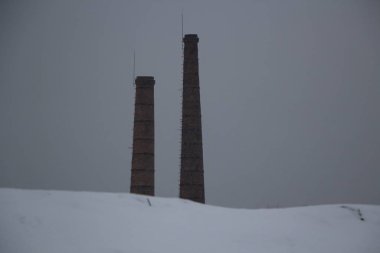 Gri gökyüzünün arka planında bir bitkinin boruları. Tuğla borular bir tepenin arkasından dikizleniyor. Sanayi bölgesi. Kışın bulutlu hava. Borulardan hava emisyonu. Asgari arkaplan.