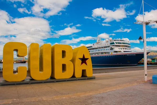 La inscripción de la CUBA. Enormes letras en el paseo marítimo, contra un cielo azul con nubes en Santiago de Cuba. Un barco turístico en el fondo . — Foto de Stock