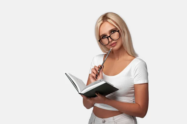 Gündelik tişört giyen genç sarışın bir kadın not defteriyle izole edilmiş beyaz arka planda dikiliyor. Mükemmel yüzlü ve gözlüklü bir kızın portresi. Model.