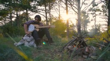 Sibirya husky köpek yakınındaki ormanda kamp ateşi ile günbatımı sırasında oturan gitar çalmak adam