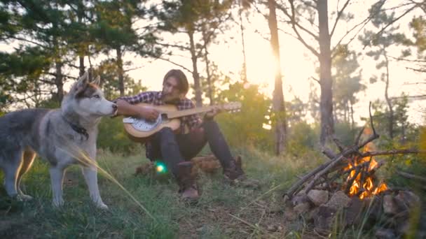 Mężczyzna Gra Gitarze Siedząc Psem Husky Syberyjski Pobliżu Ogniska Lesie — Wideo stockowe