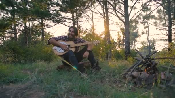 日没時に森でキャンプファイヤーの近くにシベリアン ハスキー犬と一緒に座ってギターを弾く男 — ストック動画