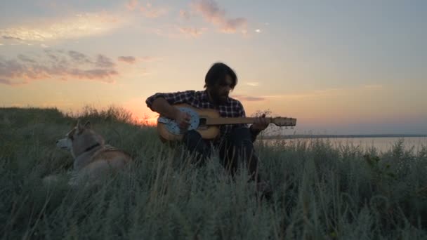 夕日の光で湖の近くにシベリアン ハスキー犬とギターを弾く男します — ストック動画