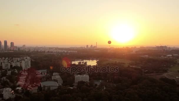 Vista aérea do horizonte de Tel Aviv ao pôr-do-sol - silhueta de edifícios, um balão de ar quente voando no céu e barcos no lago em Hayarkon Park . — Vídeo de Stock