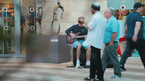 Jerusalén, Israel - 11 de mayo de 2017: Joven músico callejero guitarrista toca música cerca de monedas de dinero en la caja de la guitarra en frente de la gente transeúntes en Mamilla calle comercial centro comercial al aire libre en Jerusalén — Vídeo de stock