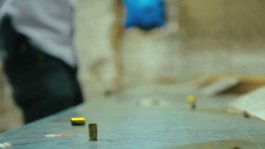 Boş tabanca mermileri atış poligonunda yere düşüp tahta masaya çarpıyor. Arka planda 9mm 'lik silahla adam ateş ediyor. Kapat, seçici odaklan ve boşluğu kopyala.