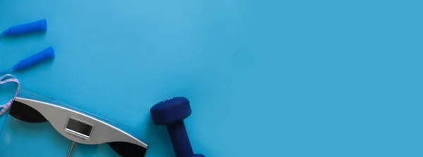 Woman measures her weight by standing her feet in socks on scale on a blue background with skipping rope and dumbbells. Flat lay composition. Healthy active lifestyle