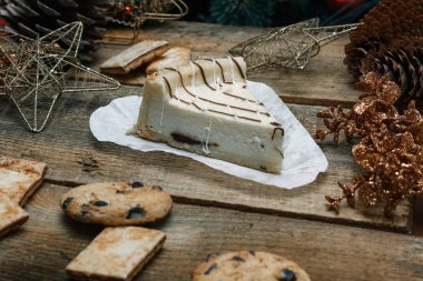 Ahşap arka plan üzerinde beyaz kağıt üzerinde güzel ev yapımı pasta peynirli kek. Sonraki çikolatalı kurabiye, Noel hediyeleri, yılbaşı ağacı, koni ve oyuncaklar. Yılbaşı kutlamak için gebe.