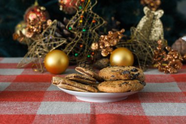 Damalı bir masa örtüsü ahşap arka plan üzerinde beyaz bir tabağa çikolatalı çerezleri. Çevre koniler ve tarçın sopa, kabaca. Yeni yıl tatili Tema
