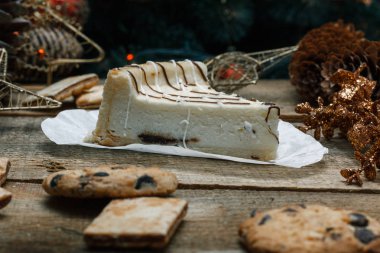 Ahşap arka plan üzerinde beyaz kağıt üzerinde güzel ev yapımı pasta peynirli kek. Sonraki çikolatalı kurabiye, Noel hediyeleri, yılbaşı ağacı, koni ve oyuncaklar. Yılbaşı kutlamak için gebe.