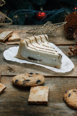 Ahşap arka plan üzerinde beyaz kağıt üzerinde güzel ev yapımı pasta peynirli kek. Sonraki çikolatalı kurabiye, Noel hediyeleri, yılbaşı ağacı, koni ve oyuncaklar. Yılbaşı kutlamak için gebe.