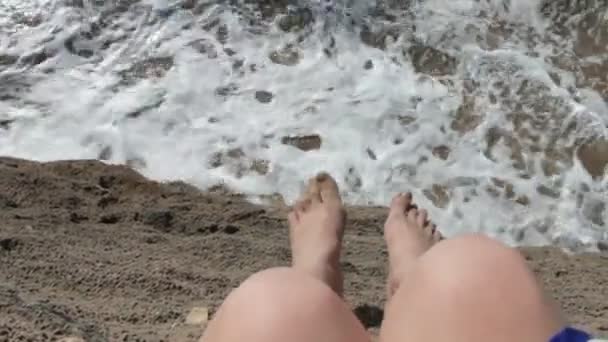 Una chica está sentada en un acantilado cerca del océano. Las piernas cuelgan. El vestido se desarrolla en el viento. Las piernas de una mujer contra el mar del océano de olas . — Vídeo de stock