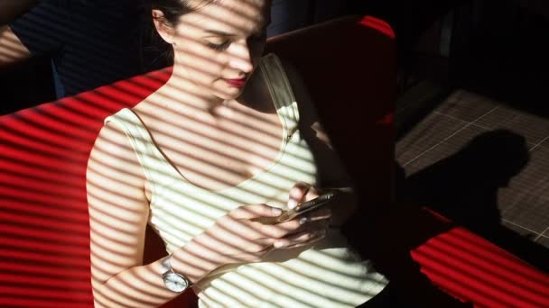 Young woman in a shirt sitting at table in cafe near window and type message on mobile phone — Stock Video