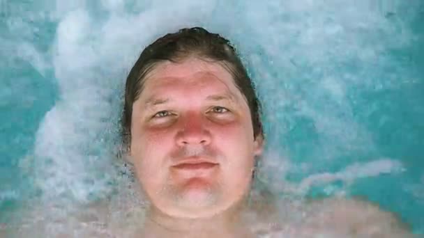 L'uomo è sdraiato si rilassa nella vasca idromassaggio e guarda la telecamera. L'uomo caucasico che riposa in una piscina giace sull'acqua. Vista dall'alto — Video Stock