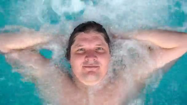 De man ligt ontspant in de jacuzzi en kijkt naar de camera. Kaukasische man rust in een pool ligt op het water. Van bovenaf bekijken — Stockvideo