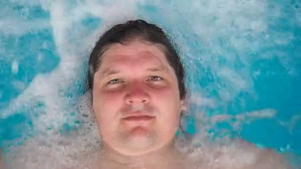 Человек лежит релаксации в джакузи и смотрит на камеру. Белый мужчина, отдыхающий в бассейне, лежит на воде. Взгляд со стороны — стоковое видео