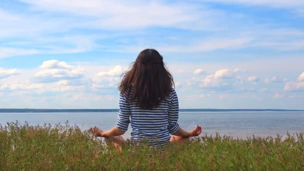 Young woman from back view in striped clothes sitting in the lotus pose on the beach of river. Caucasian woman feeling unity with nature and recreation while practicing yoga and meditating in the — Stock Video