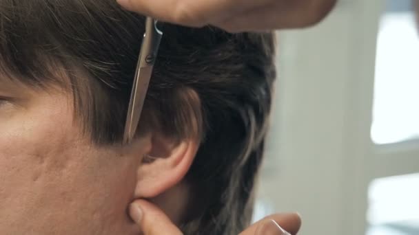 Friseur schneidet dem Kunden in einem Salon Haare an Schläfe und Ohr. Mann sitzt im Friseursessel, während er im Friseurladen mit der Schere schert. — Stockvideo