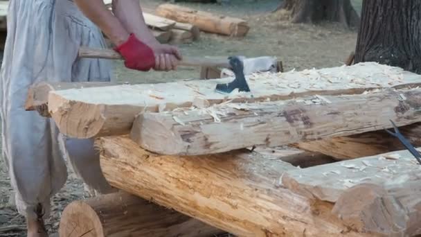 Timmerman in middeleeuwse katoenen kleding werkend met hardhout met een bijl. Een man snijdt handmatig een houten plank met een bijl. Bouwplaats. Ax heeft een log close-up gemaakt. Slivers vliegen in verschillende richtingen — Stockvideo