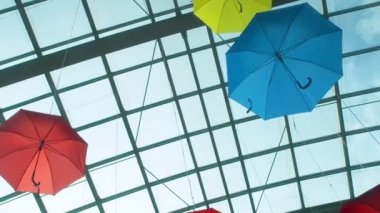Many bright colored umbrellas suspend under a transparent glass ceiling in shopping center bottom view. Beautiful multi-colored umbrellas hang under the roof of a shopping center. Colorful abstract