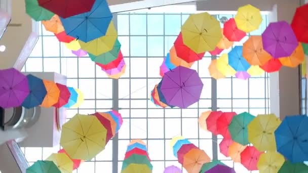 Many bright colored umbrellas spinning under a transparent glass ceiling in shopping center bottom view. Multi-colored umbrellas hang under the roof of a shopping center. Camera rotation around — Stock Video