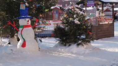 Aydınlatılmış dekoratif bir kardan adam gündüz vakti karlı bir caddenin ortasında bir Noel ağacının yanında duruyor. İngilizceye çevrilmiş mor bir tabakta çubuğa lolipop yazılıdır.