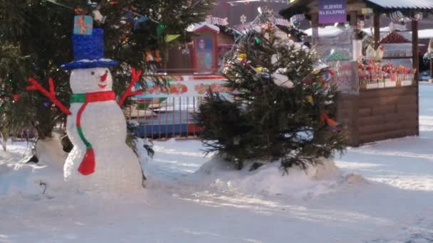 En dekorativ snögubbe med belysning står nära en julgran mitt på en snöig gata på dagen. På en lila platta i översättning till engelska skrivs Lollipop på en pinne — Stockvideo