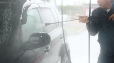 Bir adam siyah bir arabayı yıkar. Otomobil yıkama işleminin yavaş çekim videosu, oto yıkama jeti, yüksek basınçlı yıkama ve araba kiri. Yan Görünüm. Köpüklü Deterjan Kanalları