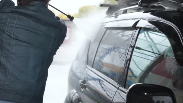 Ein Mann wäscht ein schwarzes Auto. Zeitlupenvideo einer Autowäsche an einer Selbstbedienungswaschanlage. Ein Wasserstrahl mit hohem Druck wäscht den Schmutz vom Auto ab. Seitenansicht. Abflüsse von geschäumtem Waschmittel — Stockvideo