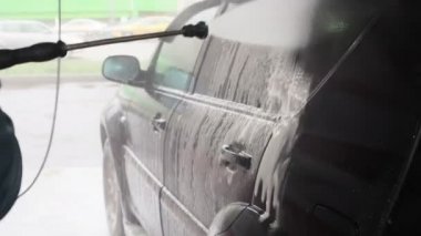 Bir adam siyah bir arabayı yıkar. Otomobil yıkama işleminin yavaş çekim videosu, oto yıkama jeti, yüksek basınçlı yıkama ve araba kiri. Yan Görünüm. Köpüklü Deterjan Kanalları