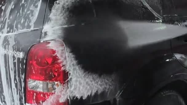 Чоловік миє чорну машину. Повільне відео руху процесу миття автомобілів на самообслуговуванні автомийки. струмінь води з високим тиском змиває бруд з автомобіля. Вид збоку. спінений миючий засіб — стокове відео