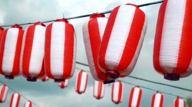 Many oriental paper red-white lanterns Chochin hanging on white cloudy blue sky background. Japanese lanterns hanging on bright sky background