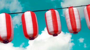 Oriental paper red-white lanterns Chochin hanging on white cloudy blue sky background. Japanese lanterns hanging on bright sky background. Pan left