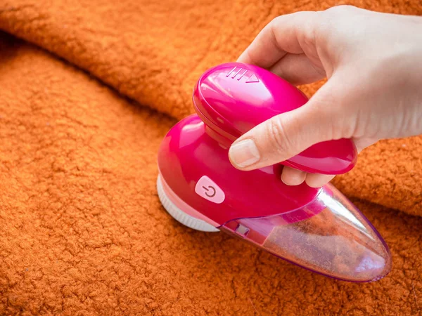 Clothes care. Lint shaver or fabric shaver or fuzz remover in female hand. Woman removing lint on orange wool coat with handheld electric defuzzer. 免版税图库照片