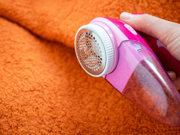 Clothes care. Lint shaver or fabric shaver or fuzz remover in female hand. Woman removing lint on orange wool coat with handheld electric defuzzer. 免版税图库图片