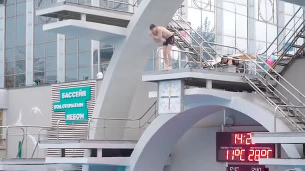 Москва, Россия - 12 октября 2019 года: Молодой человек прыгает с водонапорной башни с большой высоты в открытый бассейн. Дождь немного и холодная погода. Конкурентная платформа для дайвинга — стоковое видео