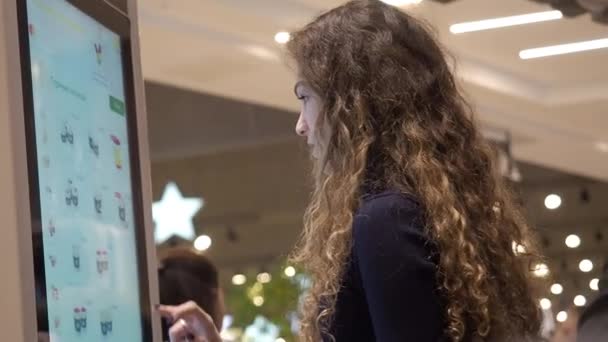 Moscou, Russie - 16 mars 2019 : Jolie fille caucasienne aux cheveux bouclés achète une boisson chaude chez McDonalds. Machines de commande libre-service à l'intérieur de McDonalds. Les gens passent devant une jeune fille achète du café en utilisant — Video