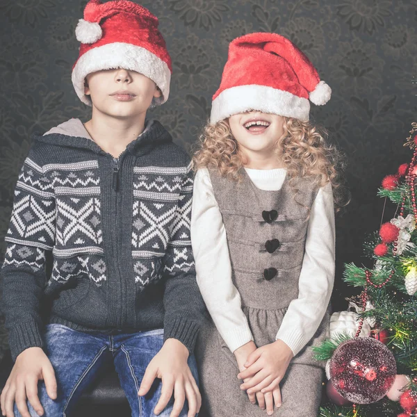 Miúdos engraçados sentados perto da árvore de Natal. Usando chapéus de Papai Noel . — Fotografia de Stock