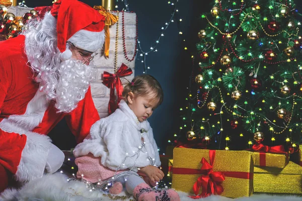 Santa en meisje speelt in de buurt van open haard, versierde kerstboom — Stockfoto