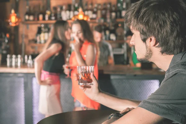 Drunk man sitting at bar, drinking cocktail, looking at girls