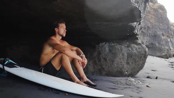 Surfer sits at cave near rocks at Bali with surfboard — Stock Video