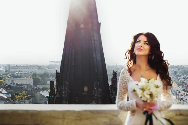 Bride daydreams with lily bouquet in her arms with cityscape on — Stock Photo, Image