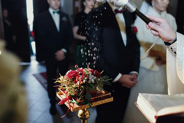 Priest sprinkles wedding rings on the red bouquet — Stock Photo, Image