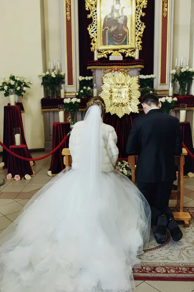 Yeni evliler dua üzerinde diz çöker kutsal kişilerin resmi içinde kilisenin önünde — Stok fotoğraf