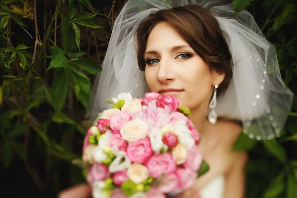 Braut steht zwischen grünen Blättern und hält einen Strauß vor ihrem Gesicht — Stockfoto