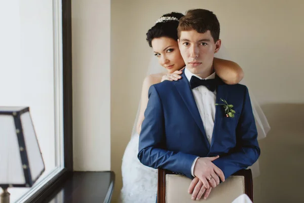 Braut steht hinter einem Bräutigam, während er sich auf den Stuhl lehnt — Stockfoto