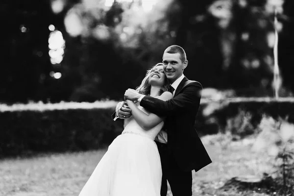 Groom hugs bride from behind while they stand in a park — Stock Photo, Image