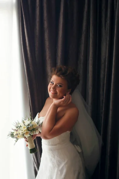 Улыбающаяся невеста отрегулирует сережку, стоящую за большим окном — стоковое фото
